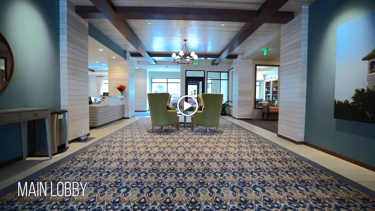 Spacious main lobby with patterned carpet, two green chairs arranged at the center, a reception desk to the left and decorative ceiling beams.