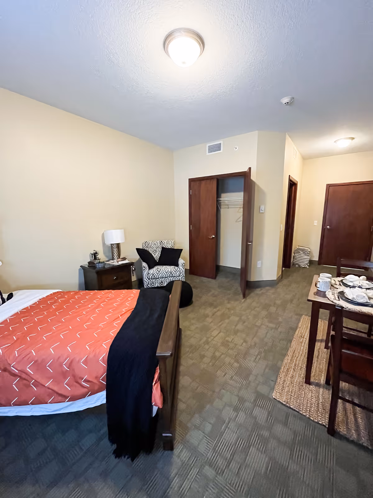 A bedroom in an assisted living facility featuring a bed with an orange and black blanket, a nightstand with a lamp and some items, a patterned armchair with two black pillows, an open closet with hangers, and a small dining table set with plates and cups on a woven rug.