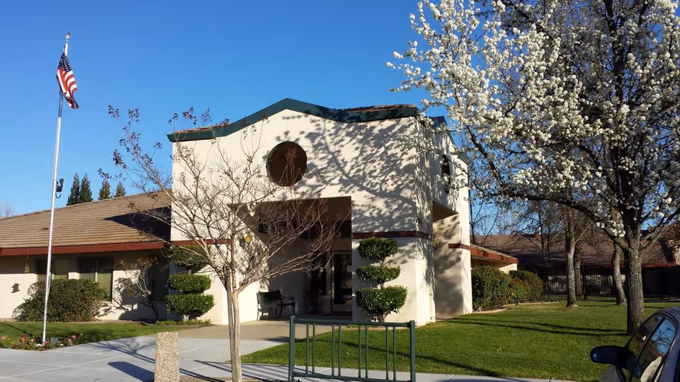 Exterior view of a single-story building with a small tower-like structure, surrounded by green grass, trees including one with white blossoms, and an American flag on a flagpole against a clear blue sky.