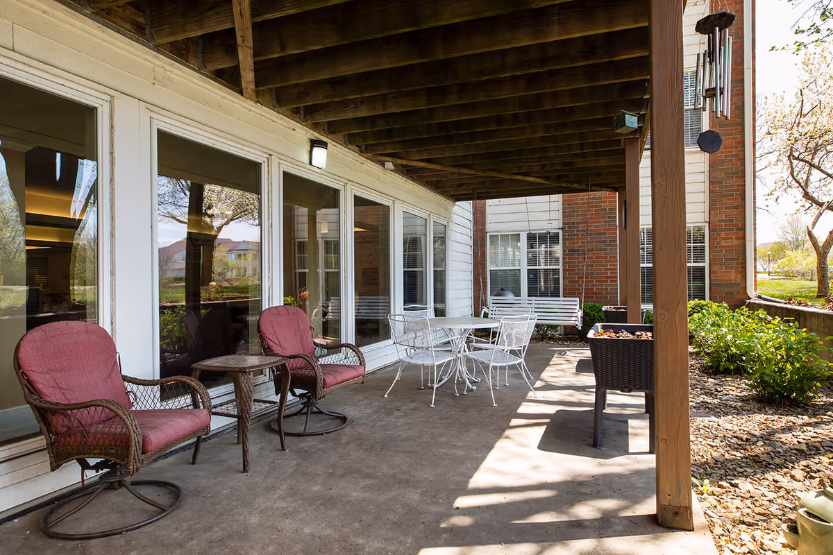 Covered outdoor patio area with two cushioned wicker chairs and a small table on the left, a white metal table with four chairs in the center, and a white porch swing in the background. The patio is attached to a building with large windows and surrounded by greenery and trees.