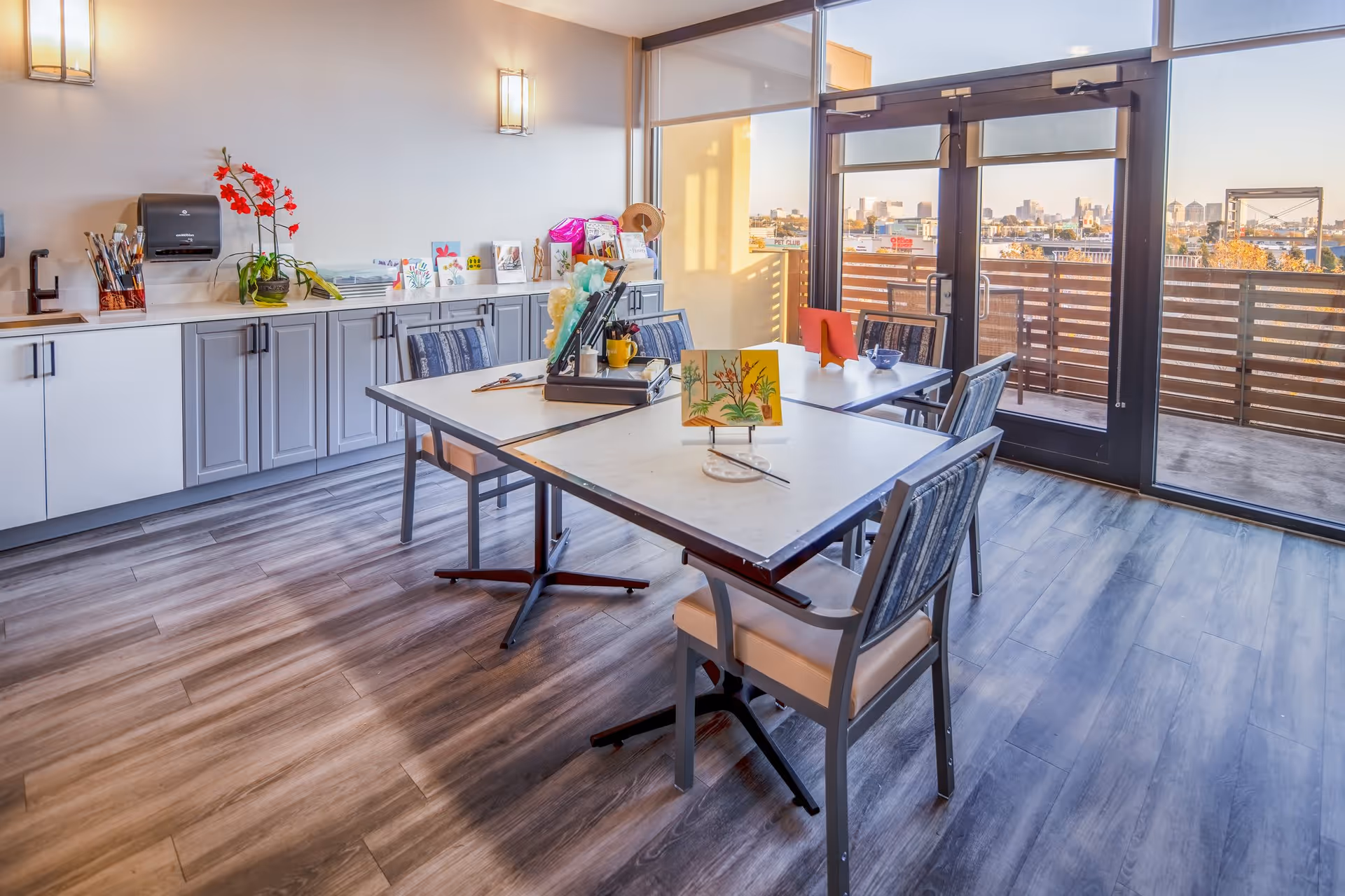 A bright and spacious activity room with tables and chairs arranged for painting and crafts. The tables have art supplies, paintbrushes, and small paintings on display. There are cabinets along one wall with additional art materials and decorations, and large glass doors open to a balcony with a city view.