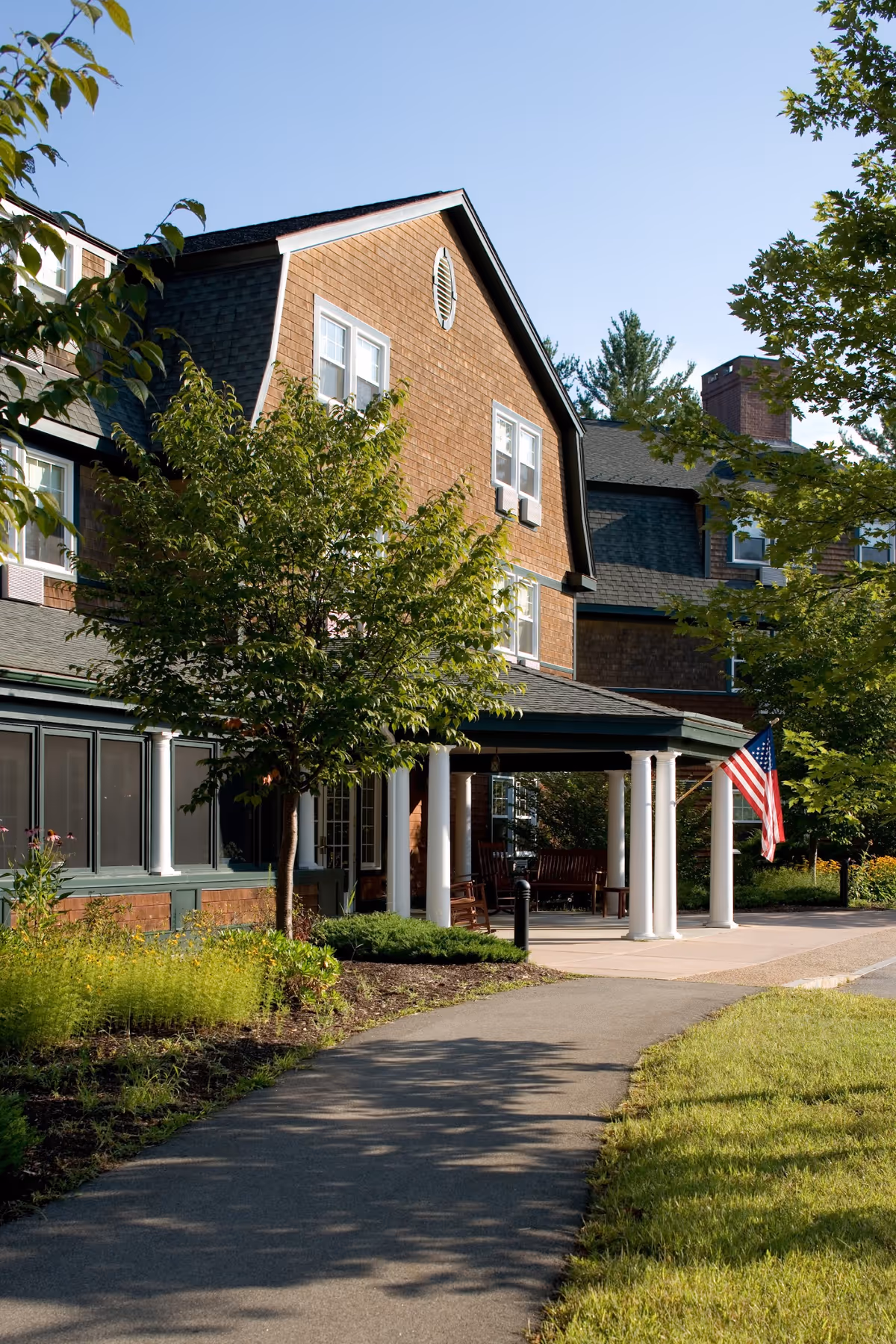 Exterior view of a senior living facility building with brown shingles, white columns supporting a covered entrance, an American flag, and surrounding greenery including trees and bushes under a clear blue sky.