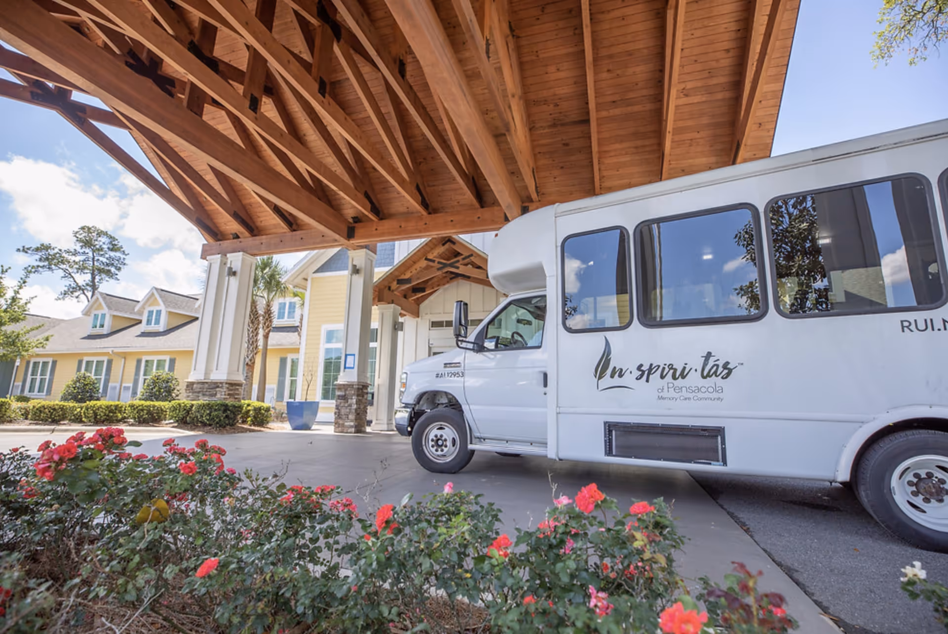 A white shuttle bus parked under a wooden canopy at the entrance of a yellow senior living facility building with white trim. There are red flowers and green bushes in the foreground, and the sky is partly cloudy.