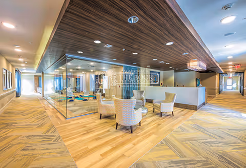A spacious and well-lit common area in a senior living facility featuring a seating arrangement with four upholstered armchairs around a small round table. The area has a mix of wood and carpet flooring, a glass-enclosed room on the left side, and a wooden ceiling panel above the seating area. Hallways extend to the right and left with multiple doors and windows allowing natural light.