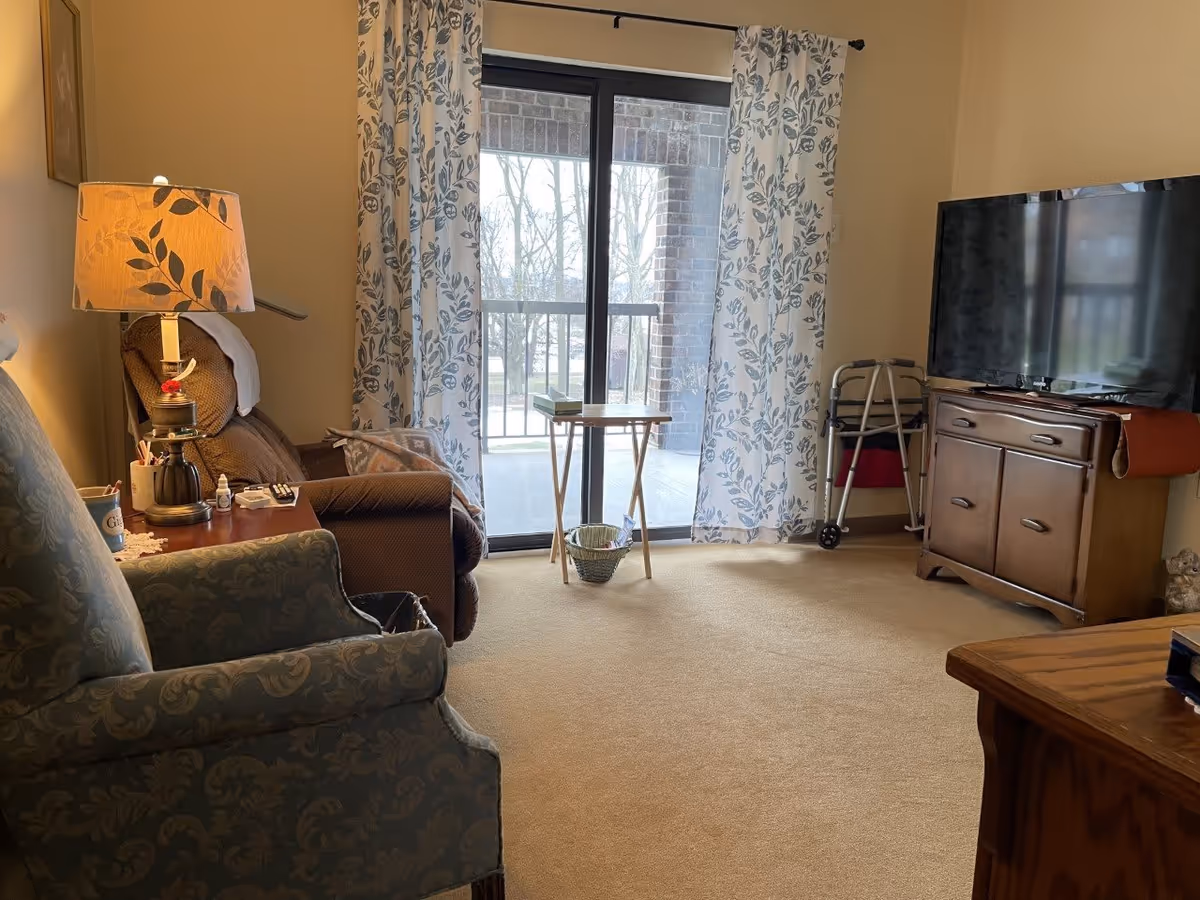 A cozy living room with a beige carpet, a blue patterned armchair, a brown recliner next to a wooden side table with a lamp, and a flat-screen TV on a wooden cabinet. Sliding glass doors with floral curtains lead to a balcony with a view of trees outside. A walker is positioned near the TV cabinet.