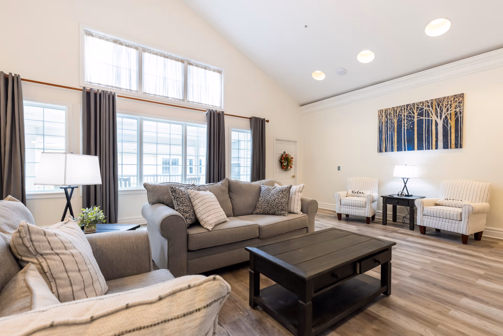 A bright and spacious living room with large windows covered by dark curtains. The room features a gray sofa with multiple patterned cushions, two striped armchairs with pillows labeled 'Relax' and 'Enjoy', a dark wooden coffee table, a side table with a lamp and plant, and a wall painting of trees. The floor is wooden, and the walls are painted white.