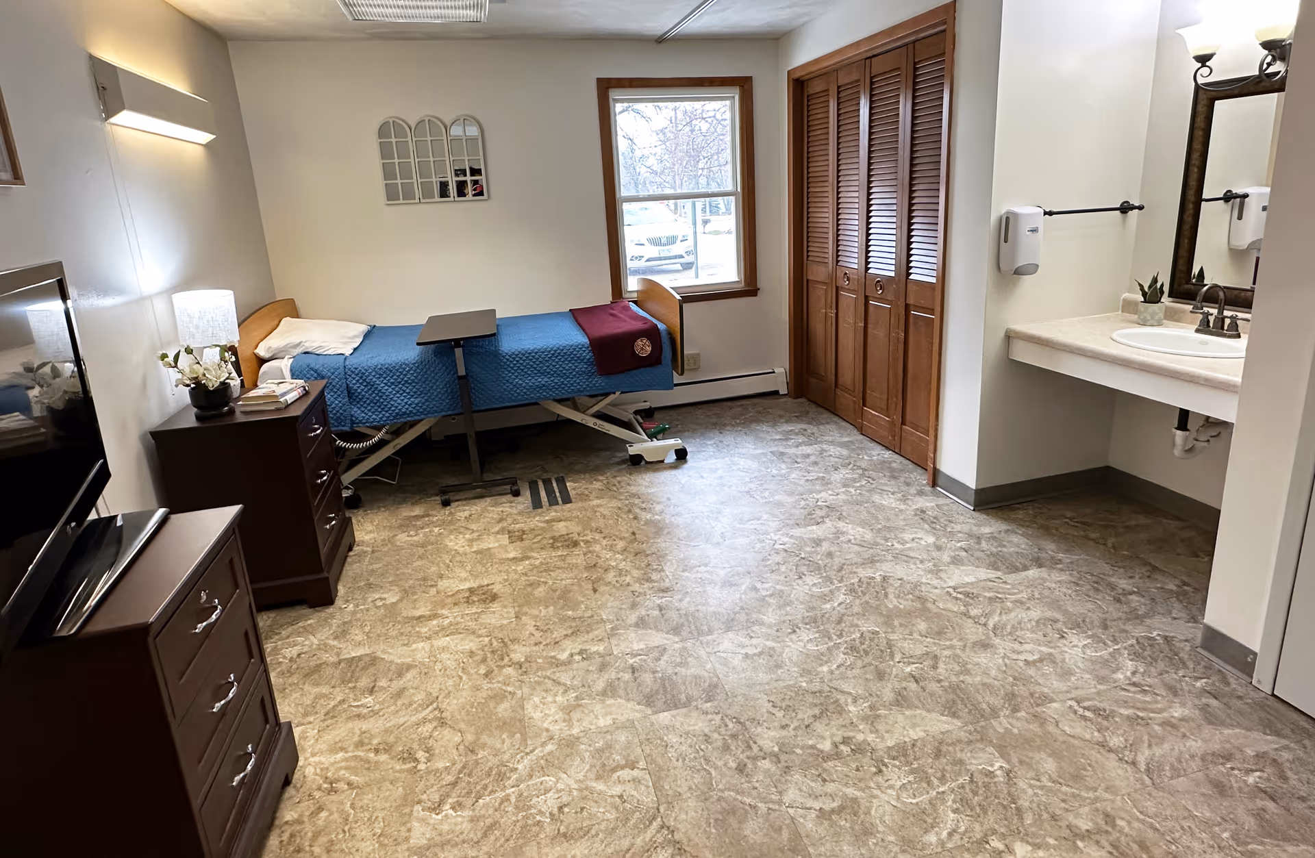 A senior care facility bedroom with a hospital-style bed covered with a blue blanket and a burgundy throw, a wooden dresser with a lamp and flowers, a TV on another dresser, a window letting in natural light, a closet with wooden louvered doors, and a sink with a mirror and soap dispenser on the right wall.