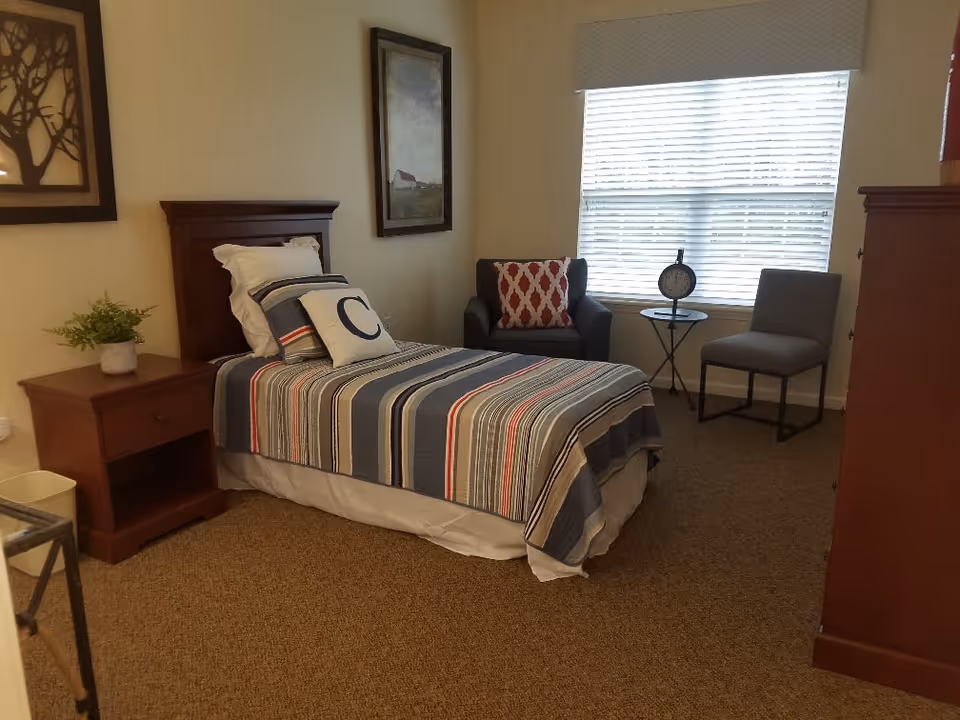 A bedroom with a single bed covered with a striped bedspread and pillows, including one with the letter 'C'. There is a wooden nightstand with a small plant, two chairs near a window with blinds, a small round table with a clock, and framed artwork on the walls.