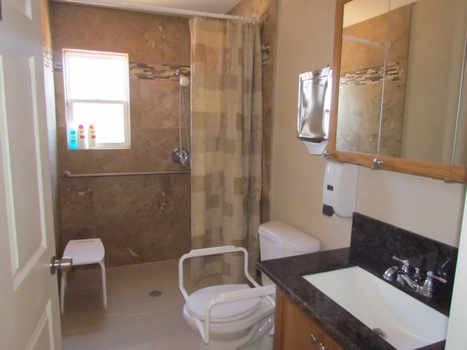 A bathroom with a walk-in shower featuring a shower curtain and a small window. There is a white shower chair inside the shower area and a toilet with white safety rails. A sink with a dark countertop and a wooden-framed mirror above it is visible. A wall-mounted soap dispenser and a paper towel dispenser are also present.