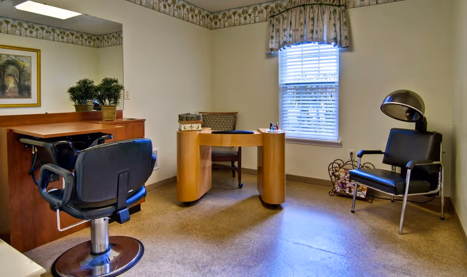 A small salon room with a black salon chair in front of a wooden counter with a mirror, a wooden manicure table with a chair, and a black hair dryer chair near a window with blinds and floral valance.
