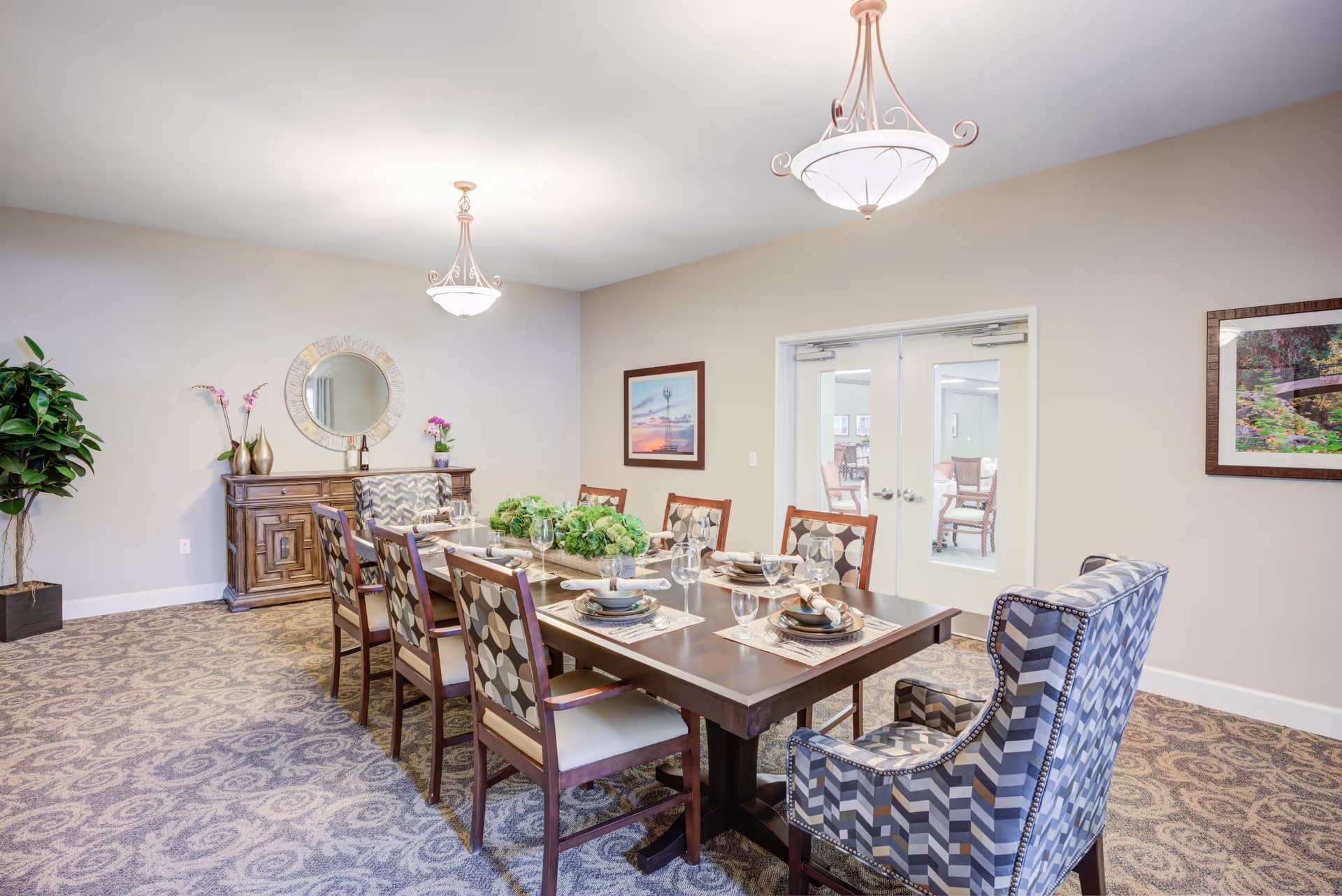 A well-lit dining room with a long wooden table set for eight people. The table is decorated with green floral centerpieces and place settings including plates, napkins, and glasses. The room features patterned carpet, two hanging light fixtures, a wooden sideboard with decorative items and a round mirror above it, a potted plant in the corner, and framed artwork on the walls. Double glass doors lead to another room with additional seating.