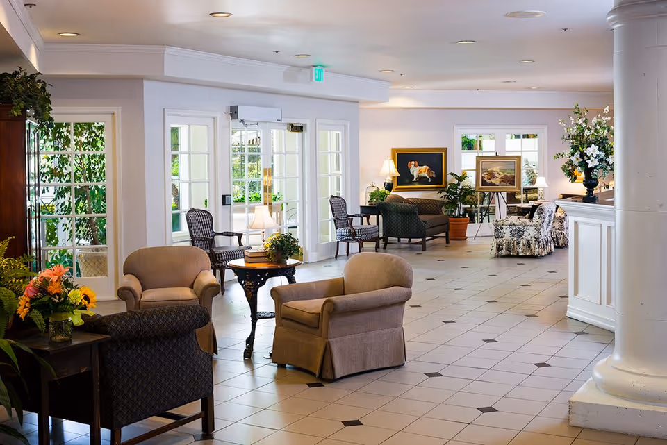 A spacious and well-lit living room area in an assisted living community featuring multiple upholstered armchairs and sofas arranged around small tables with lamps and floral decorations. The room has large windows and glass doors allowing natural light to enter, with paintings and plants adding to the cozy ambiance.
