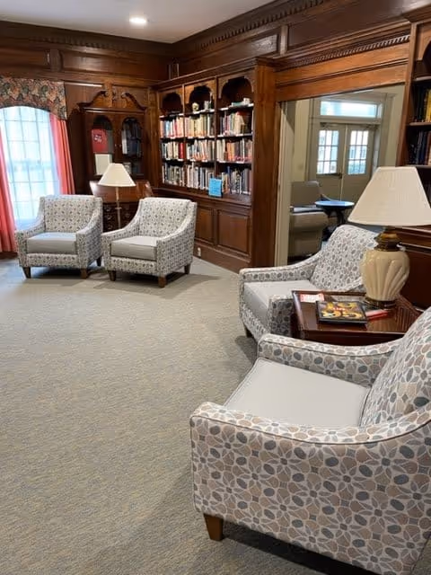 A cozy sitting area in a senior living facility with four patterned armchairs arranged around two table lamps. Behind the chairs is a wooden built-in bookshelf filled with books. The room has carpeted flooring, wood-paneled walls, and windows with floral curtains allowing natural light to enter.