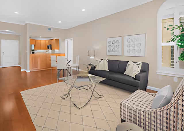 Bright open living room with a dark sofa, glass coffee table, patterned armchair and rug, with a view into the kitchen.