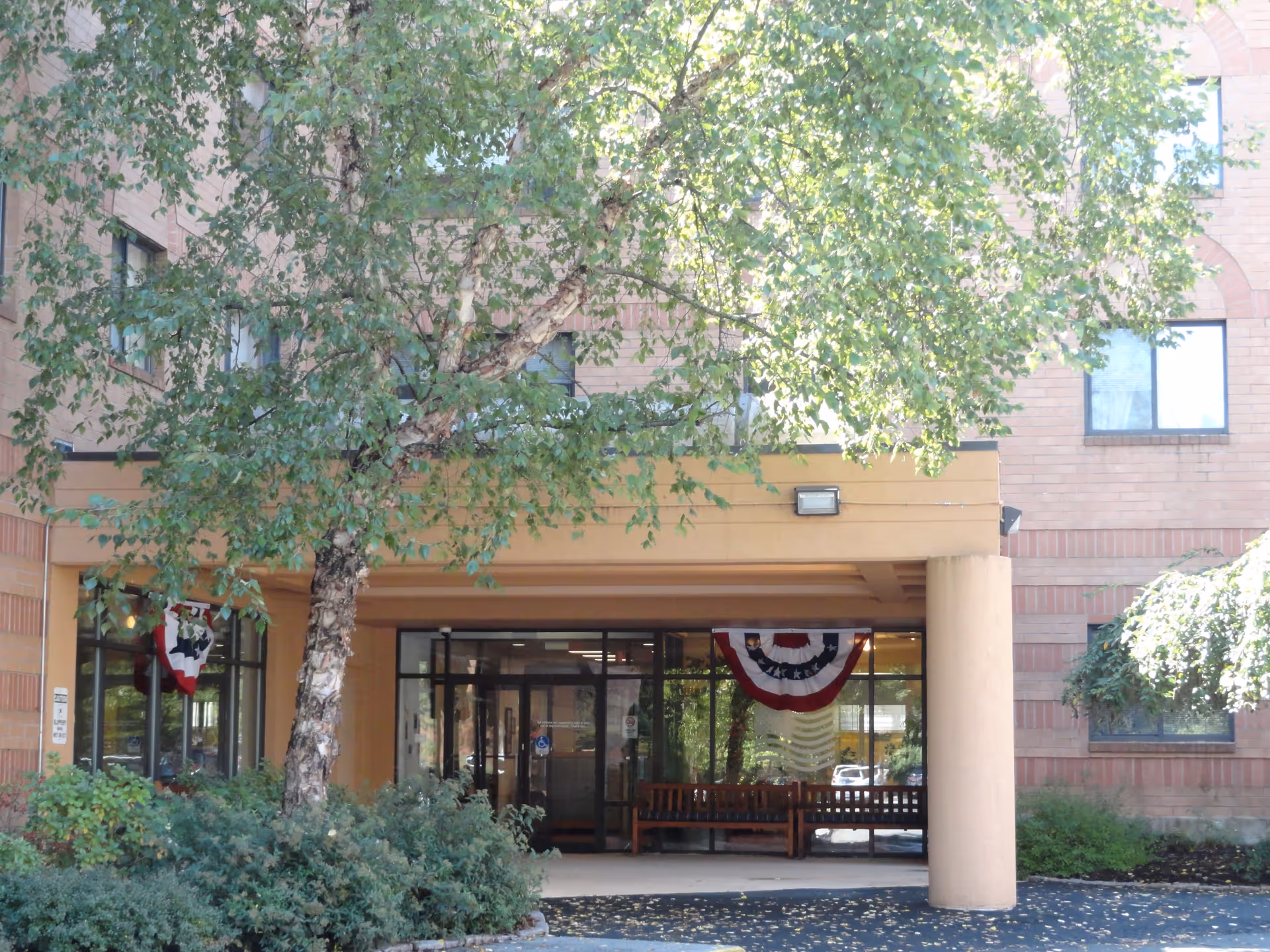 Entrance of a retirement community building with a covered drop-off area supported by columns. There are large windows and glass doors at the entrance, with patriotic red, white, and blue bunting decorations hanging inside. A tree with green leaves and some bushes are in front of the building.