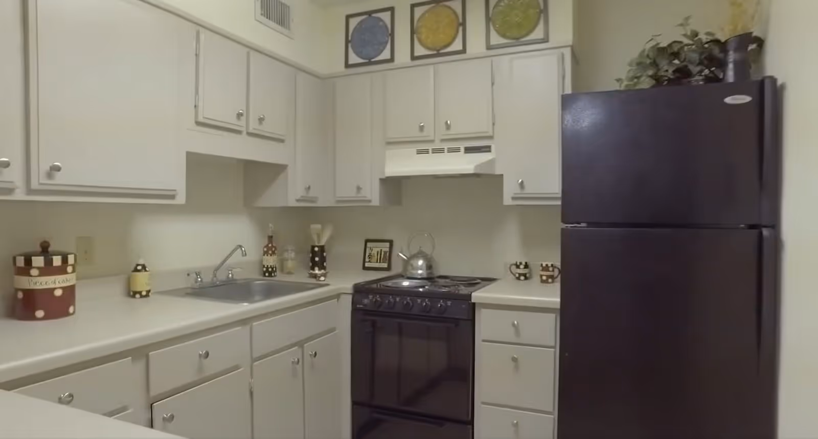 A compact kitchen with white cabinets and countertops, a black stove with a kettle on it, a black refrigerator, a stainless steel sink, and decorative items on the counters and above the cabinets.