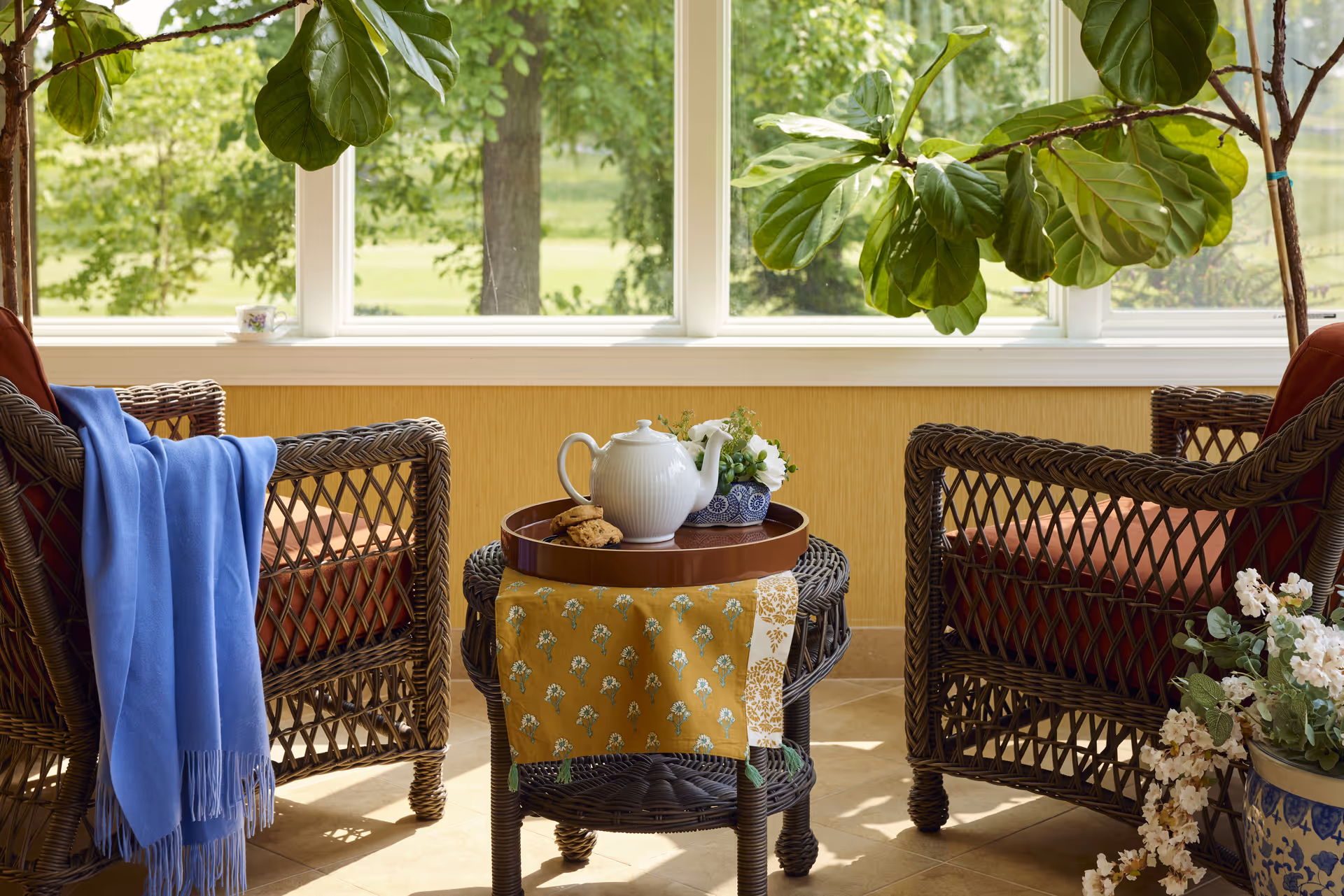 A cozy sunroom with two wicker armchairs featuring red cushions, a small wicker table between them with a yellow patterned cloth, a white teapot, cookies, and a small floral arrangement. Large windows behind the chairs show green trees and a sunny outdoor view. A blue throw blanket is draped over one chair, and potted plants with white flowers are placed nearby.