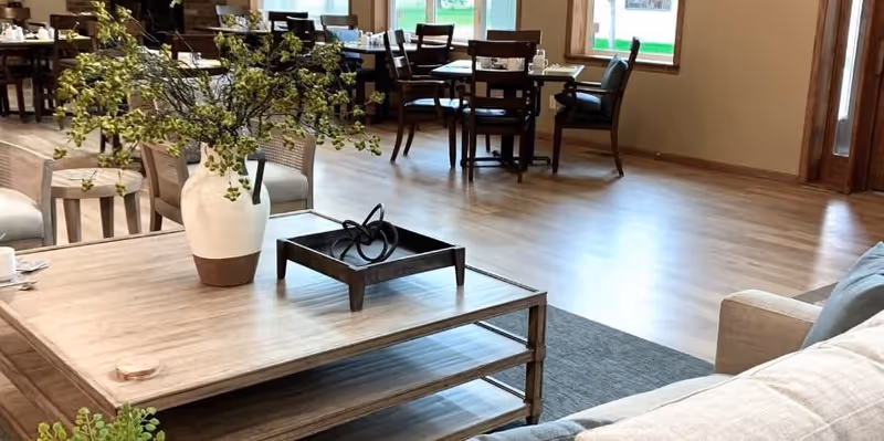 A bright and spacious common area with wooden flooring featuring multiple tables and chairs near large windows. In the foreground, there is a light-colored sofa and a wooden coffee table with a white vase containing green leafy branches and a decorative black metal sculpture.