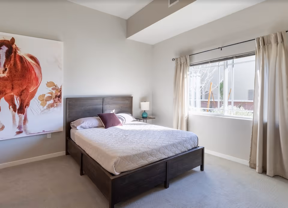 A bright bedroom with a large window covered by beige curtains allowing natural light to fill the room. The room features a dark wooden bed frame with a white quilted bedspread and two pillows, one white and one purple. A small bedside table with a lamp is next to the bed. On the wall to the left, there is a large painting of a brown horse with some plants.
