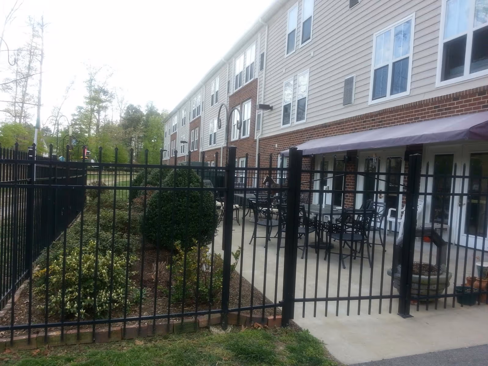 Outdoor patio area at Sandston Plateau Senior Apartments with black metal tables and chairs behind a black metal fence. The building has a brick and siding exterior with multiple windows and a purple awning above the patio doors. There are landscaped bushes and plants along the fence.