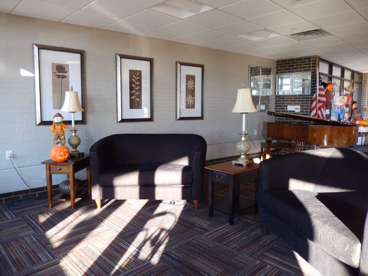 Sunlit lounge with dark upholstered sofas, side tables and lamps, framed wall art on a brick wall and a piano with an American flag in the background.