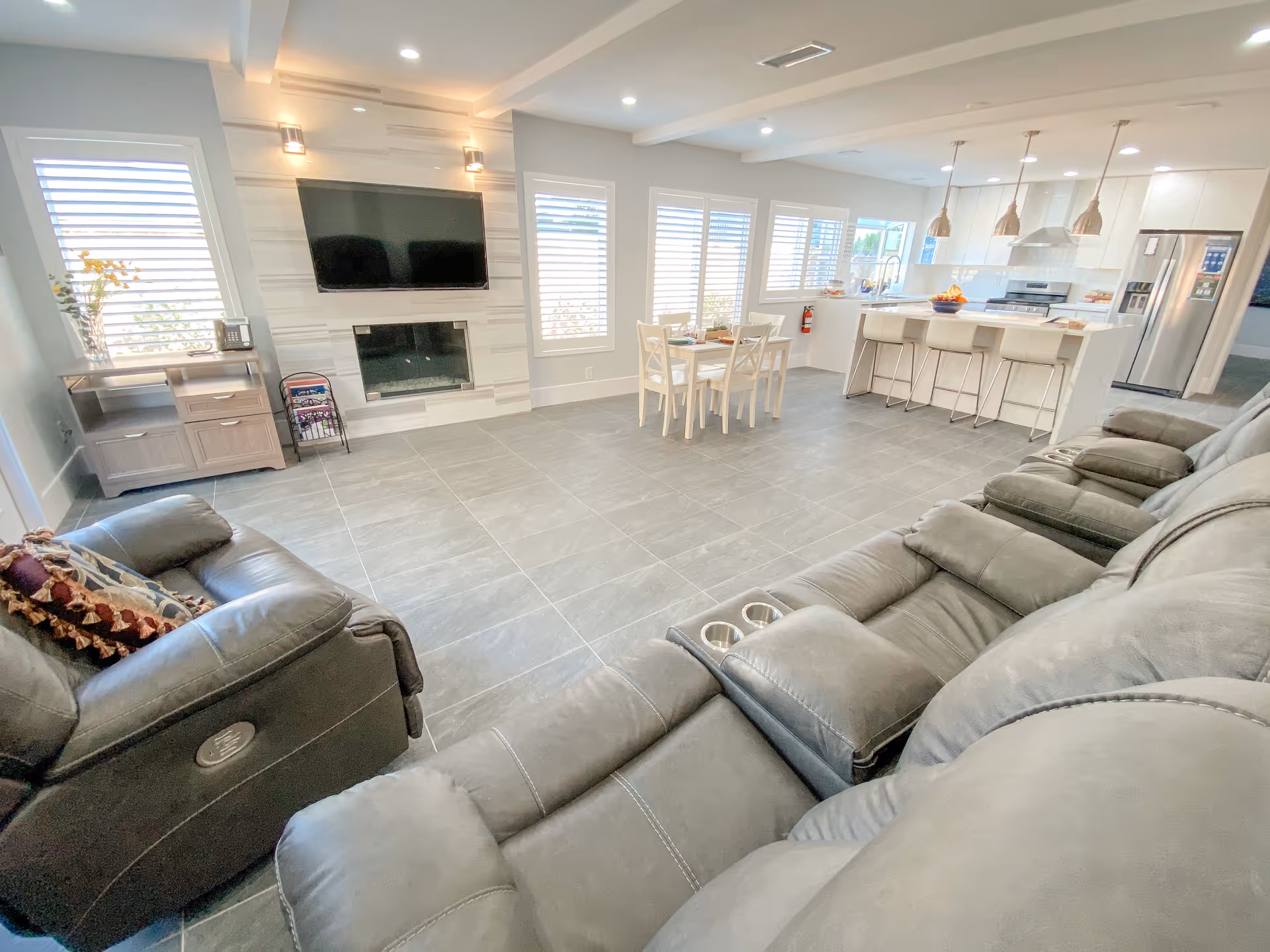Open-plan bright living room and kitchen with reclining leather sofas facing a wall-mounted TV above a fireplace, a small dining table, and a kitchen island with barstools.