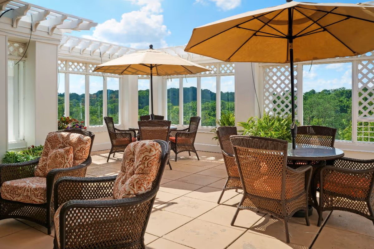 A bright and spacious outdoor patio area with wicker chairs and tables, some shaded by large beige umbrellas. The patio is surrounded by white lattice walls and large windows offering a view of green trees and a blue sky with scattered clouds.