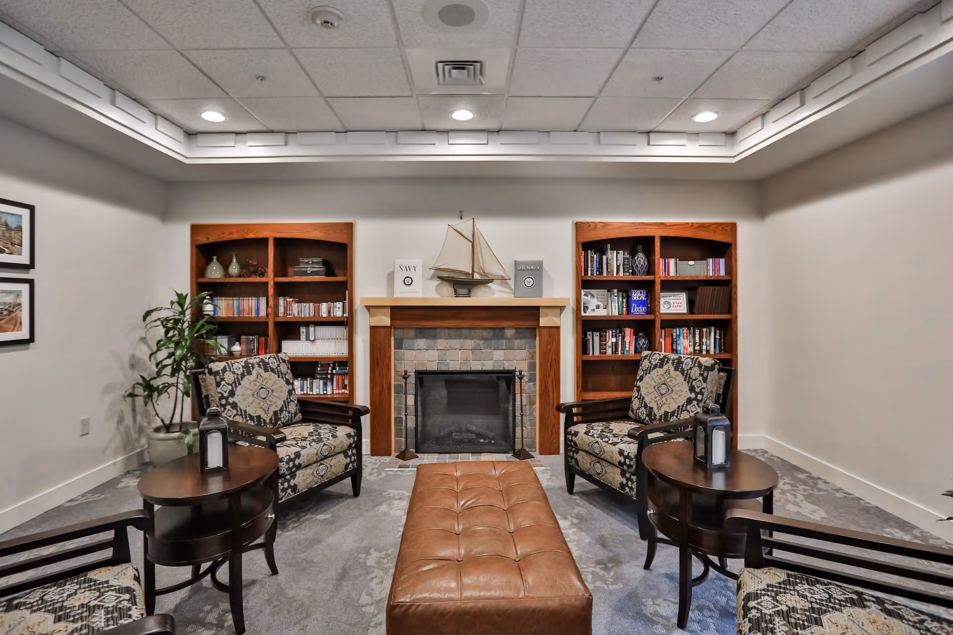 A cozy living room area with a fireplace centered between two wooden bookshelves filled with books and decorative items. Two patterned armchairs are placed on either side of the fireplace, each accompanied by a small round wooden side table with a lantern on top. A brown leather tufted bench is positioned in front of the fireplace. The room has a light gray carpet and white walls with recessed ceiling lights.