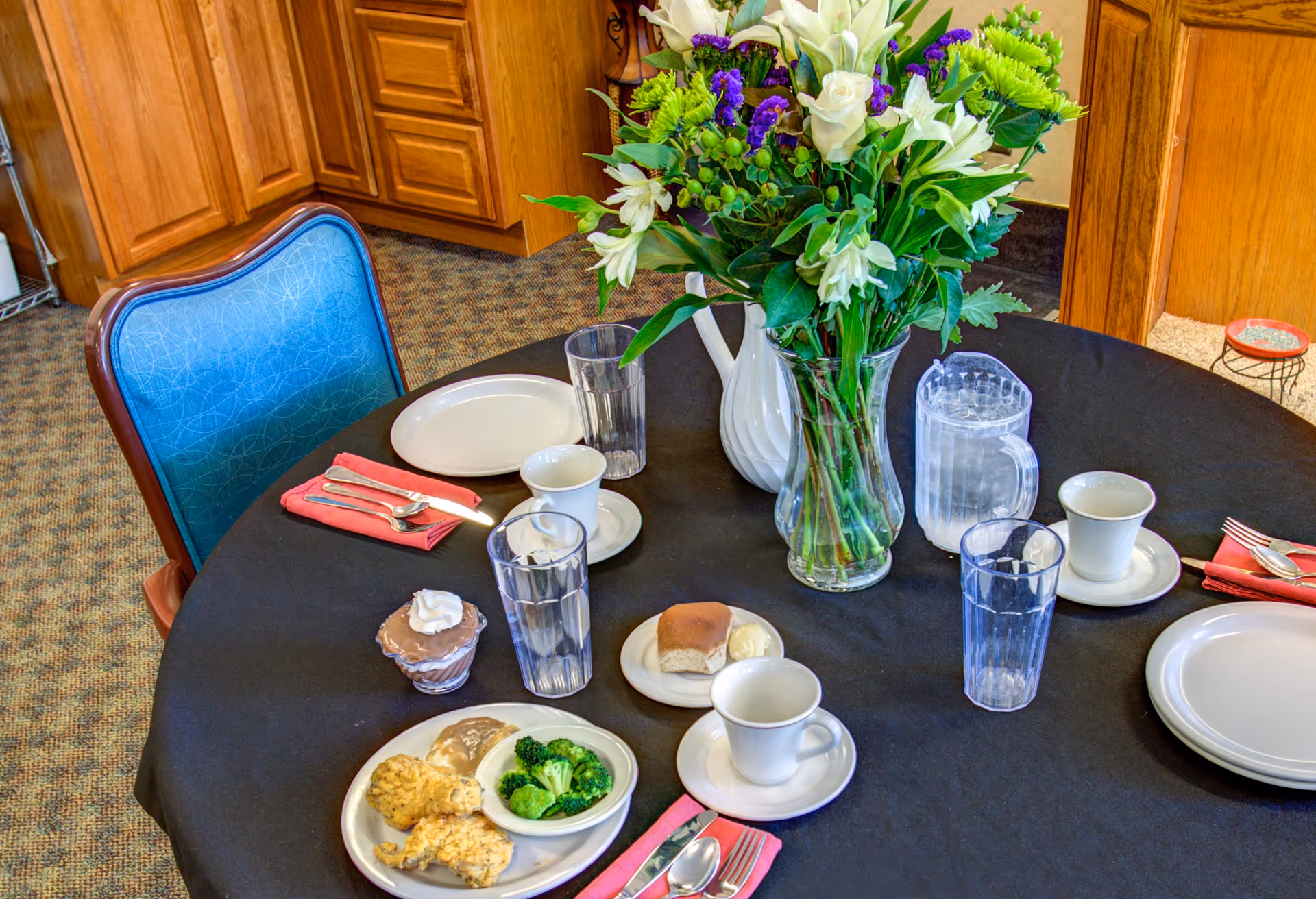 Round dining table in a communal dining room set with plates, cups, glasses, a pitcher, a vase of flowers, and small plates of food.