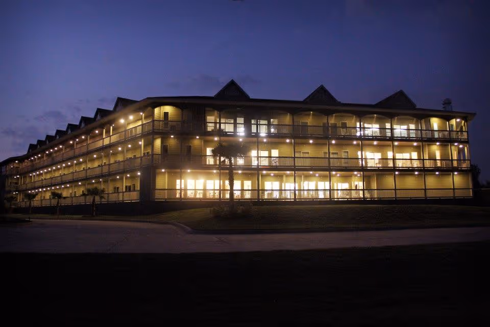 Exterior view of a three-story building at dusk with multiple balconies and bright interior lights illuminating the windows. The building has a peaked roof and is surrounded by a paved driveway and some small palm trees.