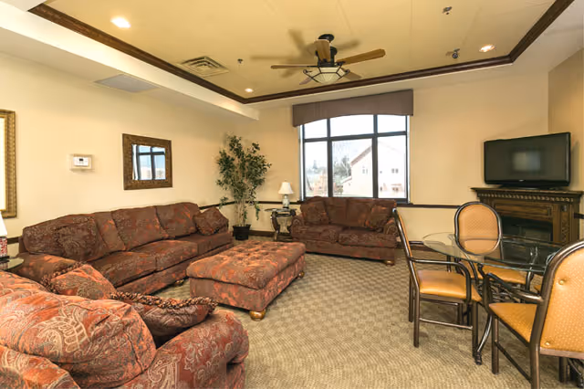 Comfortable lounge with patterned sofas and ottoman, a TV on a stand, a small dining table with chairs, and a large window.