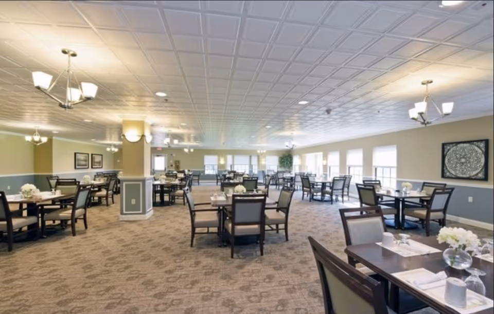 Spacious dining room with multiple tables and chairs arranged neatly. Each table is set with white napkins, glassware, and small floral centerpieces. The room has large windows allowing natural light to fill the space, beige walls with blue wainscoting, and a patterned carpet. Ceiling lights and chandeliers provide additional lighting.