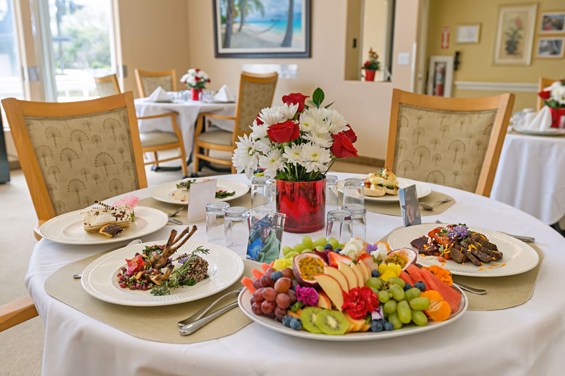 A dining table set with a white tablecloth and beige placemats, featuring a centerpiece of red and white flowers in a red vase. The table is arranged with plates of gourmet food including a fruit platter, lamb chops, steak with sauce, and dessert. Four wooden chairs with patterned upholstery surround the table. In the background, other tables with similar settings and a painting on the wall are visible in a bright dining room.