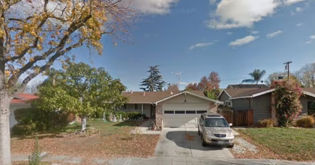 A single-story residential building with a driveway, parked car, and leafy trees along the street.