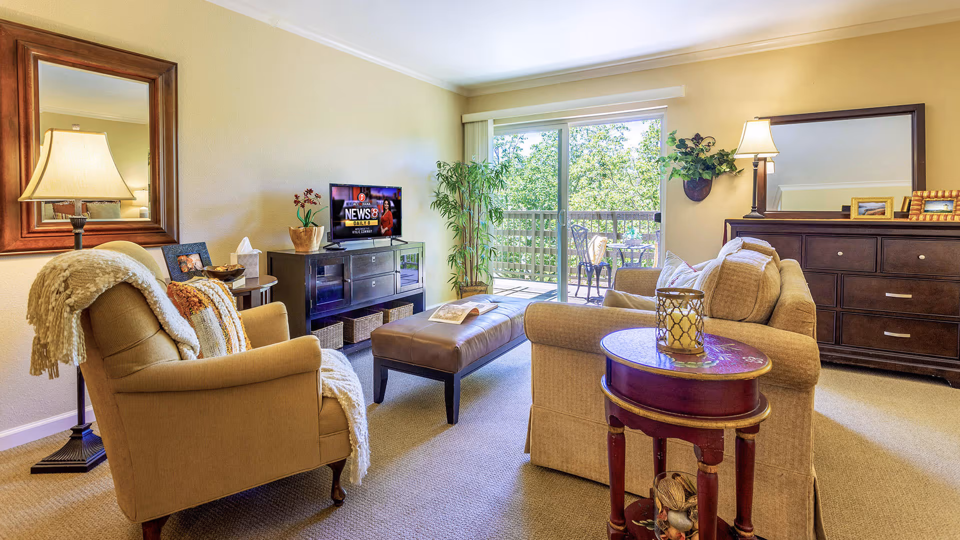 A cozy living room with beige walls and carpet, featuring a beige armchair with a throw blanket, a matching sofa, a dark wooden dresser with a large mirror, a TV on a dark wooden stand, a small round side table with a candle holder, and a sliding glass door leading to a balcony with outdoor seating and greenery visible outside.