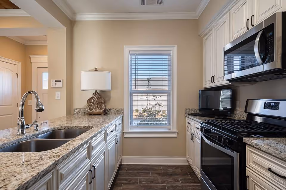 A modern kitchen with granite countertops, white cabinetry, a double sink with a chrome faucet, a stainless steel gas stove, microwave, and a small black toaster oven. There is a window with blinds in the center of the back wall and a decorative table lamp on the left countertop.