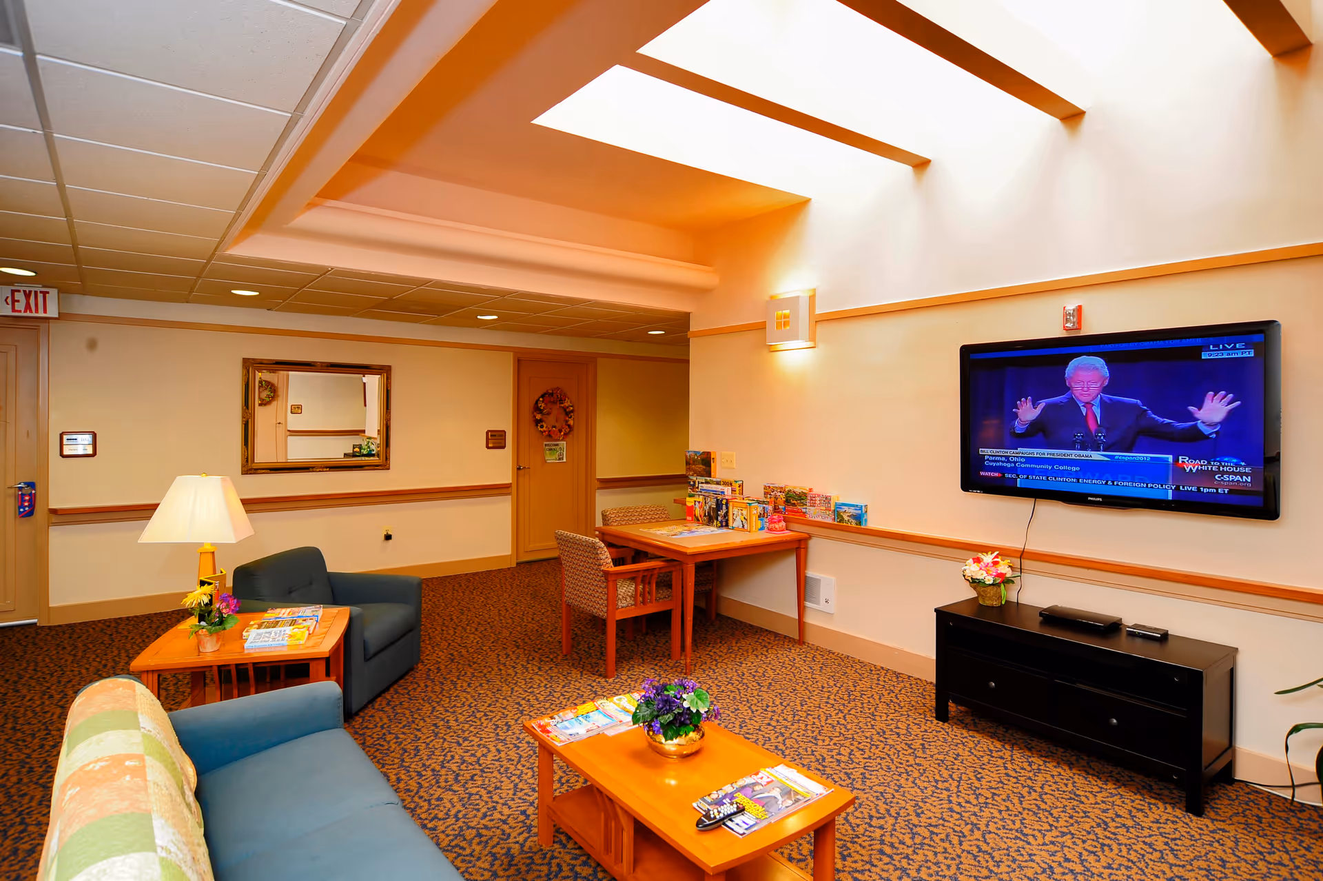 A cozy senior living community common area with a blue couch, armchair, wooden coffee and side tables with flowers and magazines, a wall-mounted flat-screen TV showing a news program, a table with chairs and board games, and warm lighting from ceiling and wall fixtures.