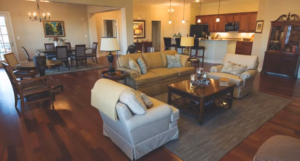 A spacious and well-lit living area featuring a beige sofa with multiple cushions, two armchairs, a wooden coffee table on a large rug, and a side table with a lamp. In the background, there is a dining area with a table and chairs under a chandelier, and an open kitchen with a breakfast bar and stools. The room has wooden flooring and warm, neutral-colored walls with framed artwork.