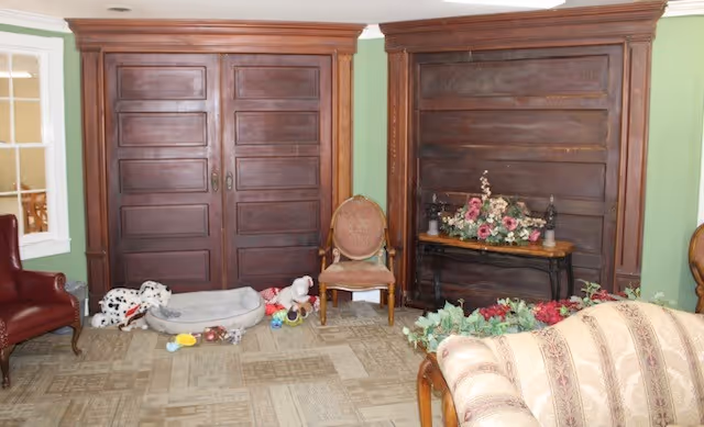 A cozy interior room with green walls and wooden paneling. The room features a patterned beige carpet, a red leather armchair, a vintage upholstered chair, a floral arrangement on a wooden table, and a beige patterned sofa. There are also several dog toys and a pet bed on the floor near the wooden panels.