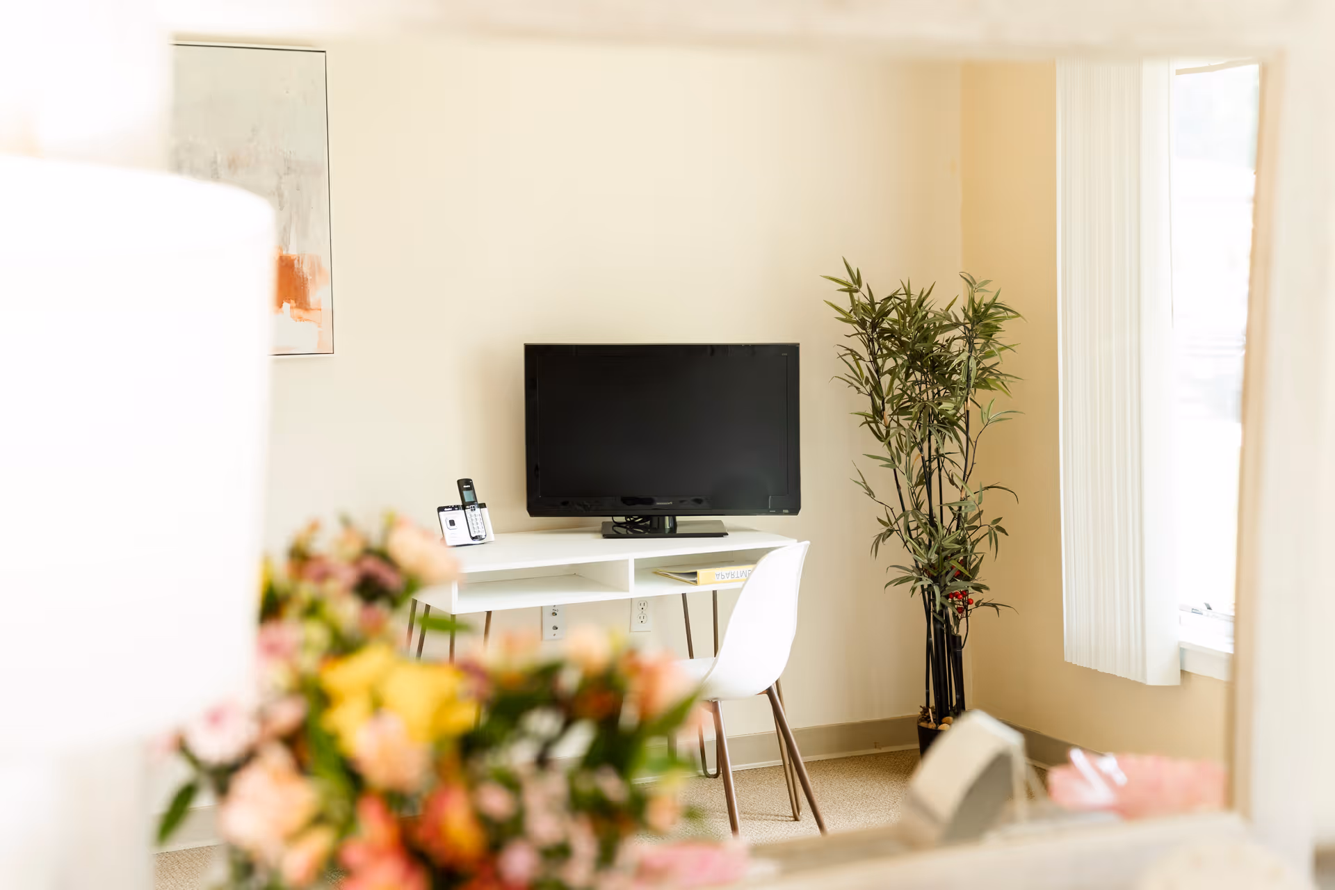 A bright room with a white desk holding a flat-screen TV and a cordless phone. A white chair is positioned at the desk. There is a tall green plant in a black pot near a window with vertical blinds. A blurred bouquet of colorful flowers is visible in the foreground.