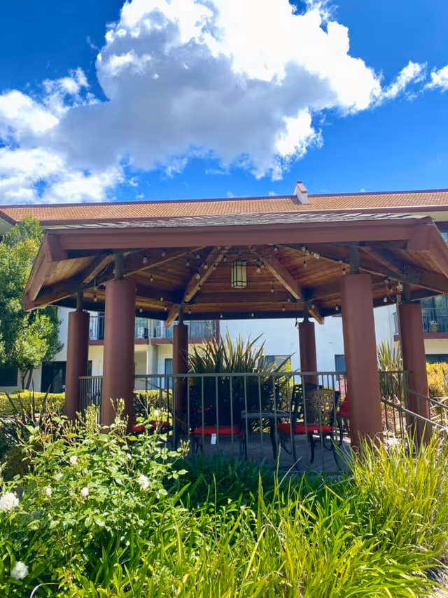 A wooden gazebo with a peaked roof supported by thick brown pillars, surrounded by lush green plants and flowers. There are chairs with red cushions inside the gazebo. In the background, part of a multi-story building with balconies is visible under a blue sky with scattered clouds.