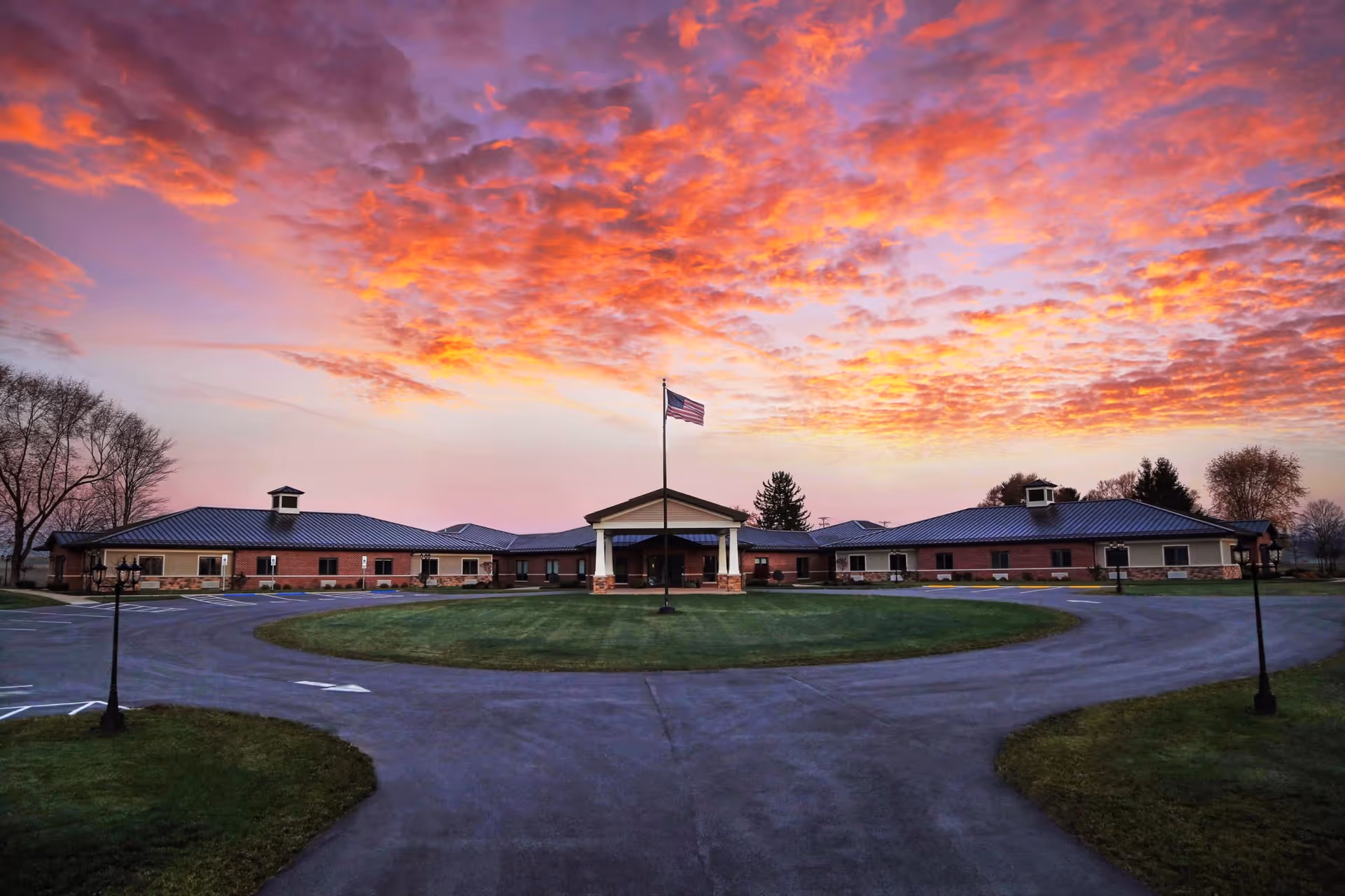 Wide exterior view of The Legacy Assisted Living Community building at sunset with a circular driveway and an American flag in the center. The sky is filled with vibrant orange and purple clouds.