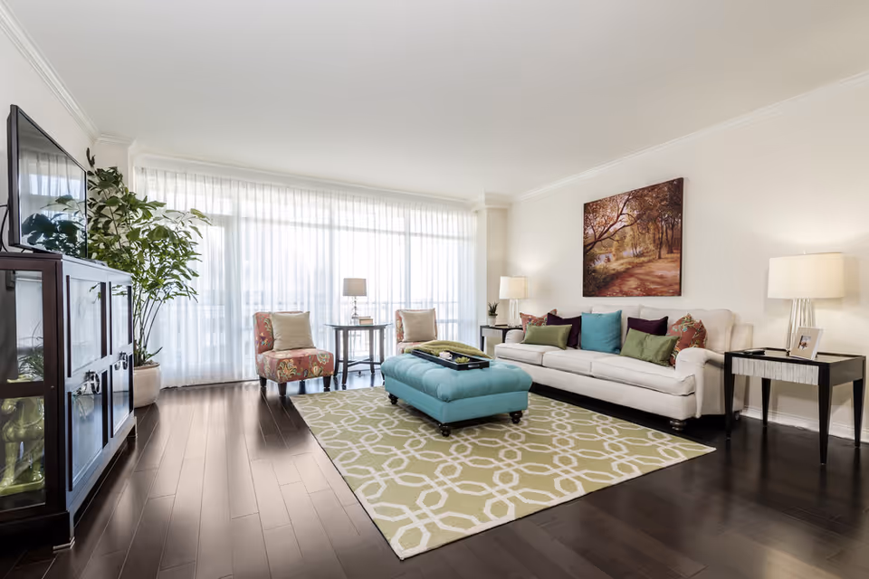 A bright living room with a white sofa and colorful pillows, a teal ottoman on a patterned rug, armchairs, TV cabinet, and a wall-length window with sheer curtains.