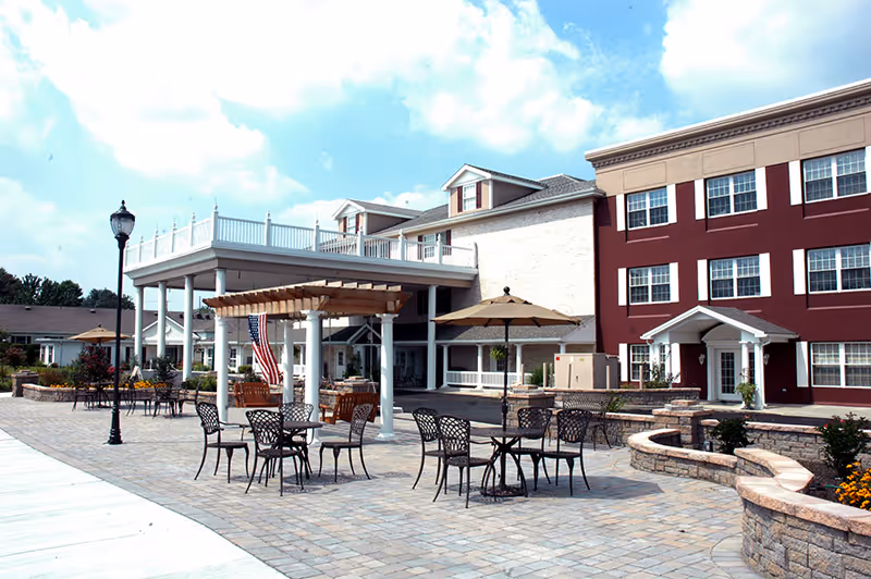 Outdoor patio area of a senior living facility with several metal tables and chairs, a pergola with an American flag, and a large umbrella providing shade. The building in the background has multiple windows and a red and beige exterior.