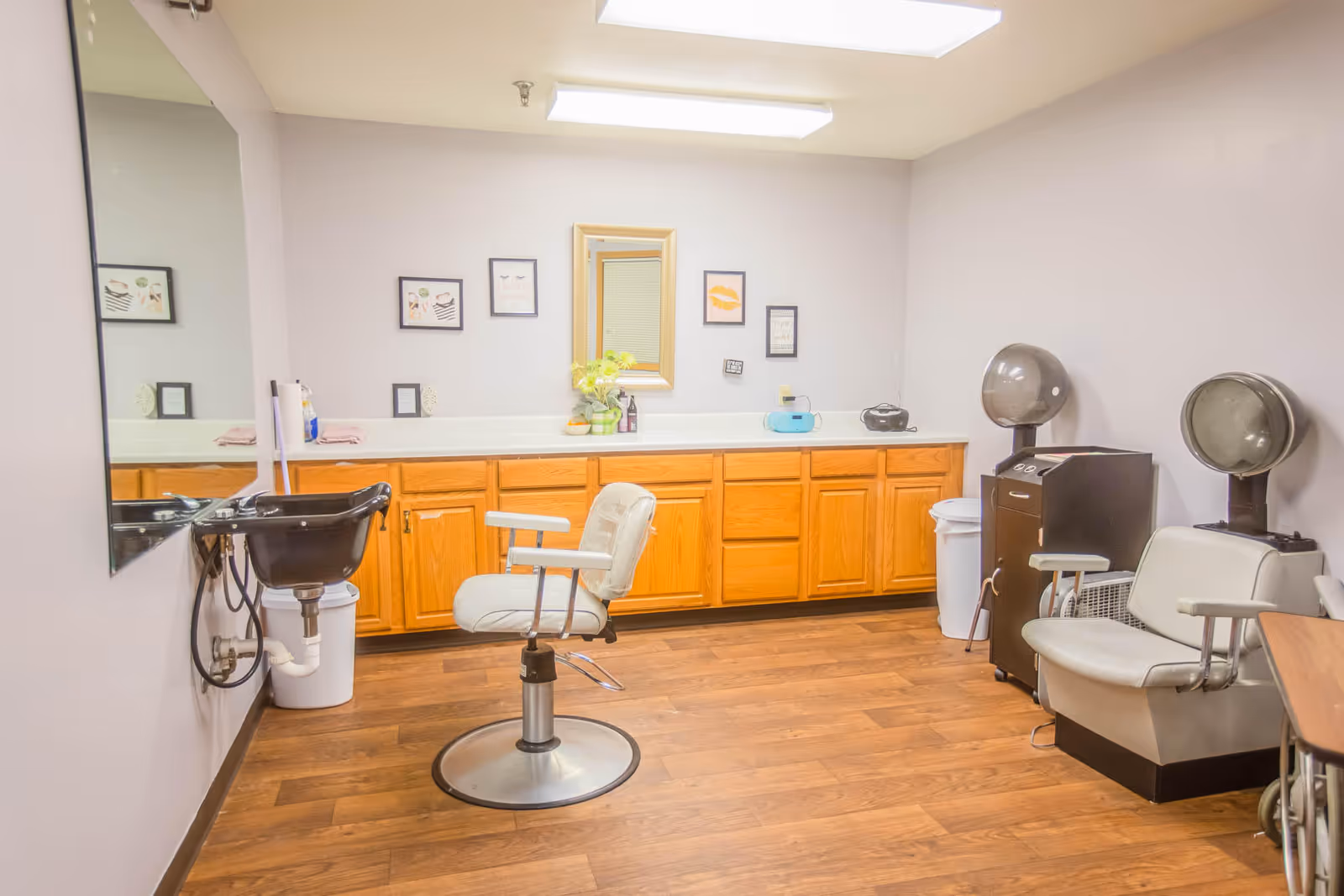 A senior living facility hair salon room with a white salon chair in front of a black wash basin on the left, wooden cabinets with a countertop and a mirror on the back wall, and two vintage hair dryers with chairs on the right side. The room has wood flooring and light-colored walls with framed pictures.