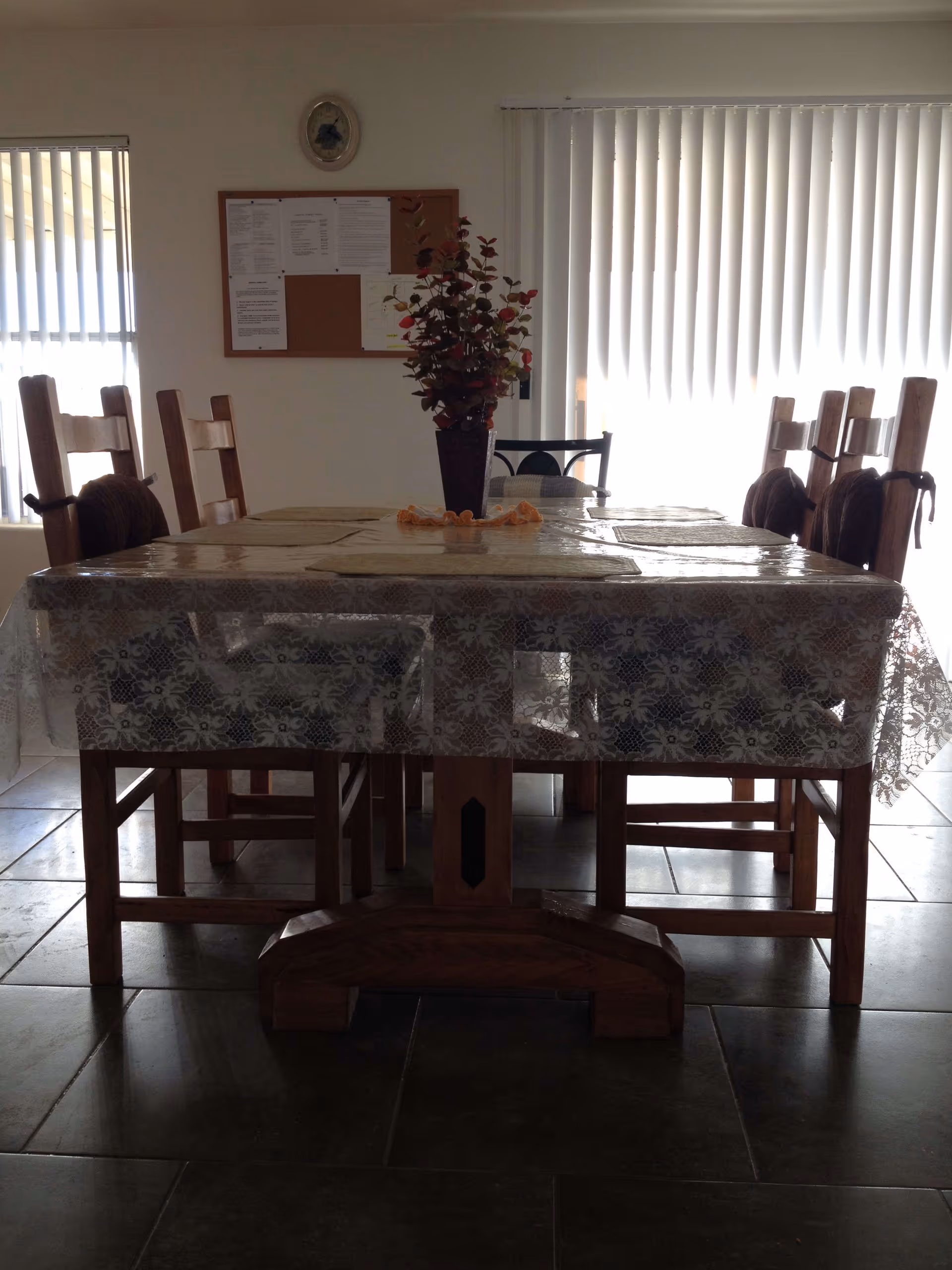 A dining room with a wooden table covered by a lace-patterned plastic tablecloth and six wooden chairs around it. A vase with artificial flowers is placed in the center of the table. The room has tiled flooring, vertical blinds on the windows, and a bulletin board on the wall.