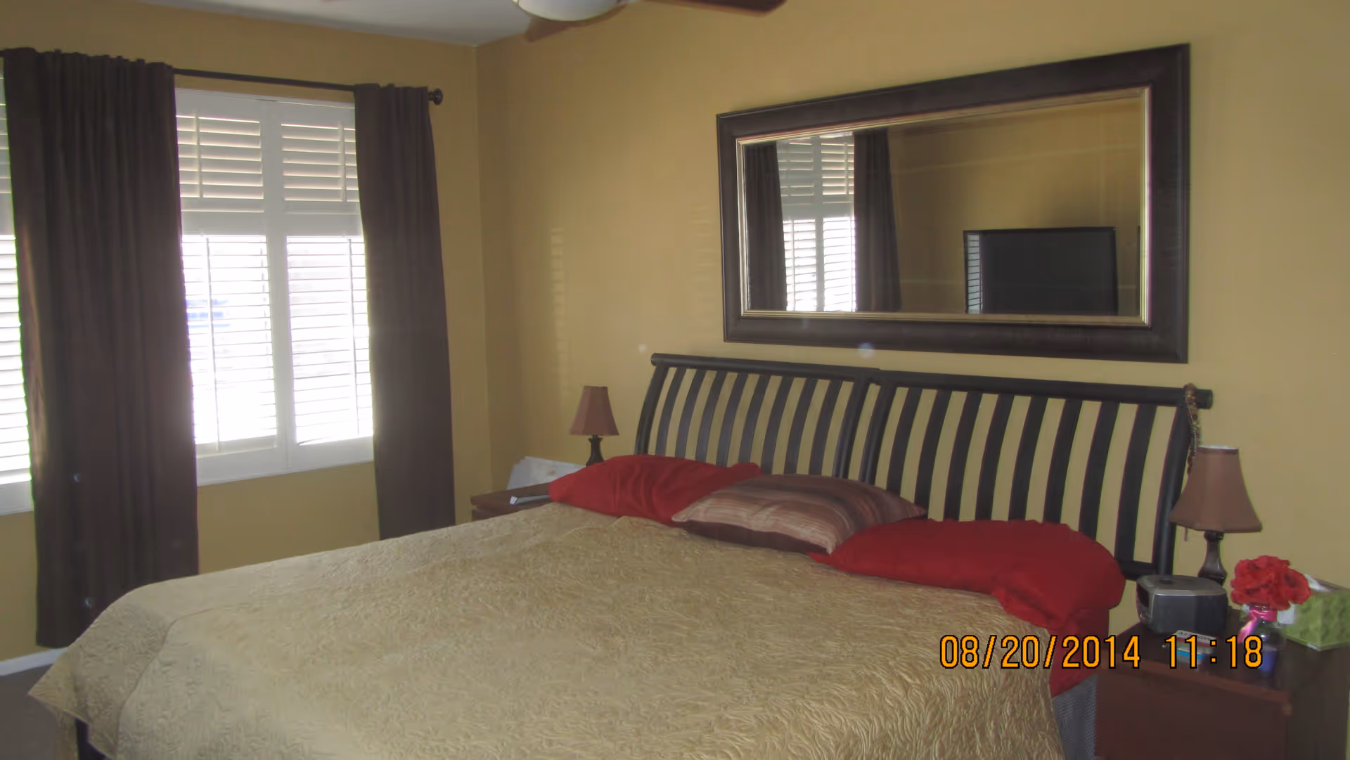 Bedroom with a large bed with red pillows, a slatted headboard beneath a wall mirror, nightstands, and a window with shutters and dark curtains.