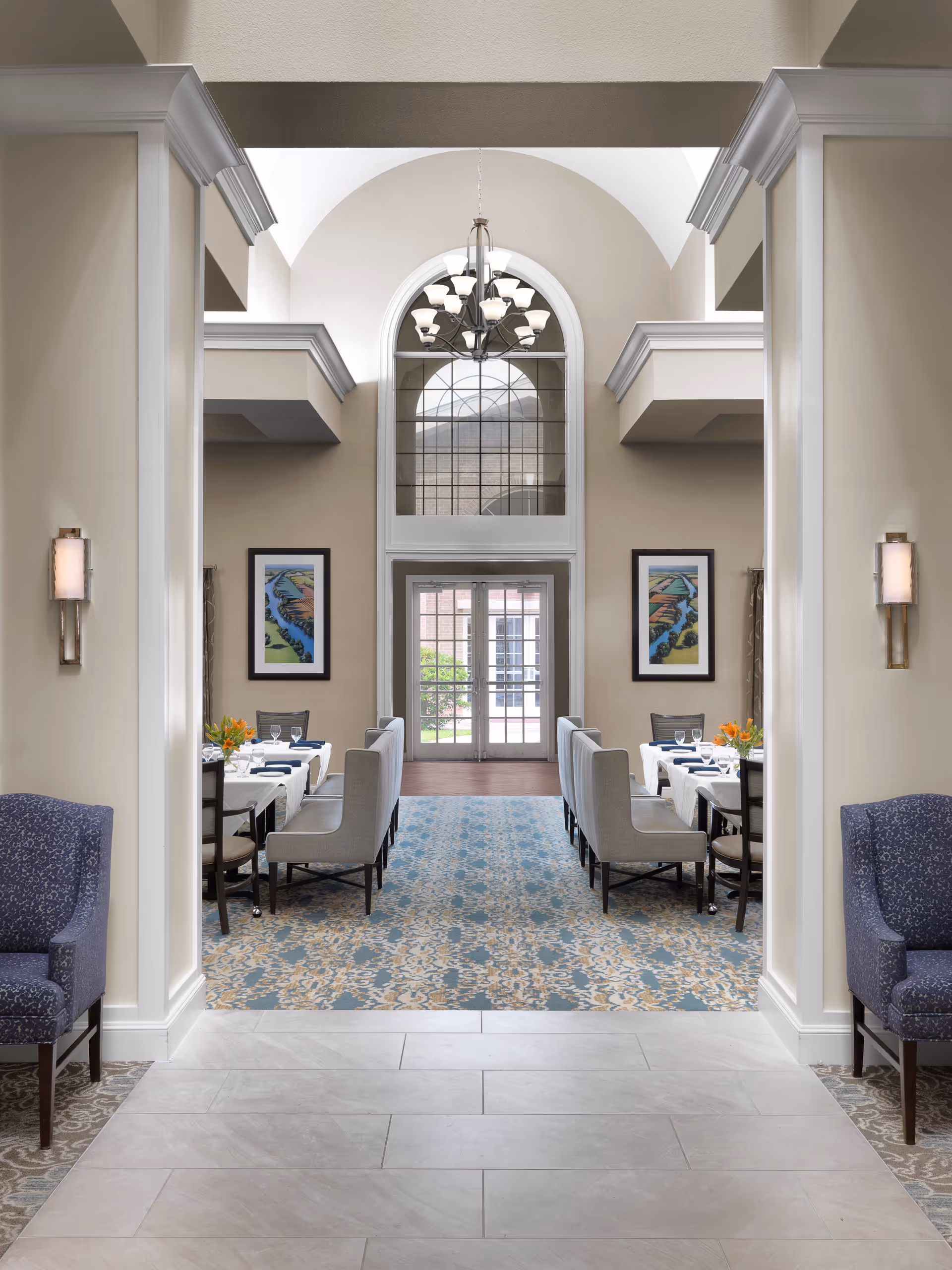 Formal dining room with a high arched window and chandelier, featuring rows of tables and upholstered chairs.