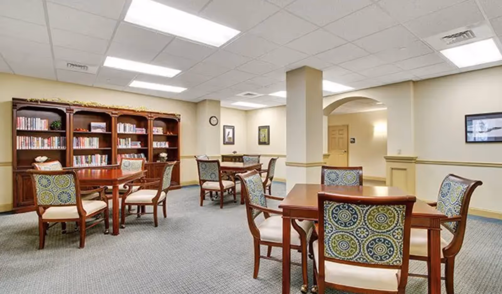 A well-lit common room with multiple wooden tables and chairs featuring patterned upholstery. There is a large wooden bookshelf filled with books along one wall, a clock, framed artwork, and a carpeted floor. The room has a neutral color scheme with beige walls and ceiling tiles with fluorescent lighting.