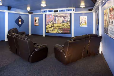 A small home theater room with blue walls and ceiling, featuring two rows of brown leather recliner chairs facing a large movie poster for 'Law for Tombstone' on the front wall. Additional movie posters are displayed on the side walls, and the room is carpeted in dark blue.